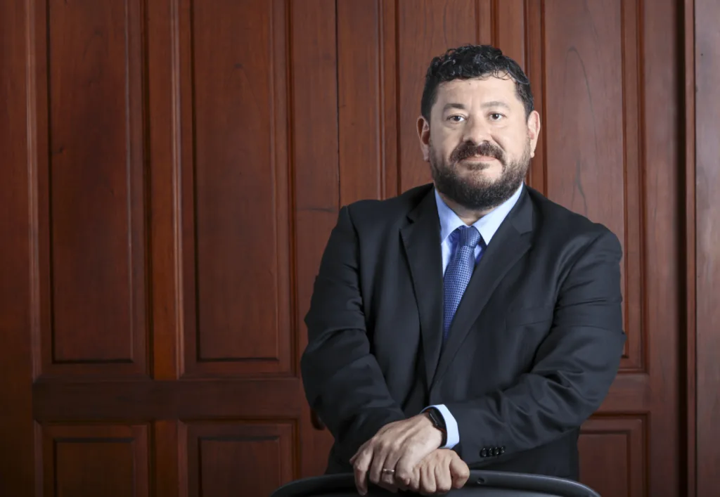 Hombre profesional con barba y bigote, vestido con traje oscuro y corbata azul, posando con las manos sobre una silla frente a una pared de madera.