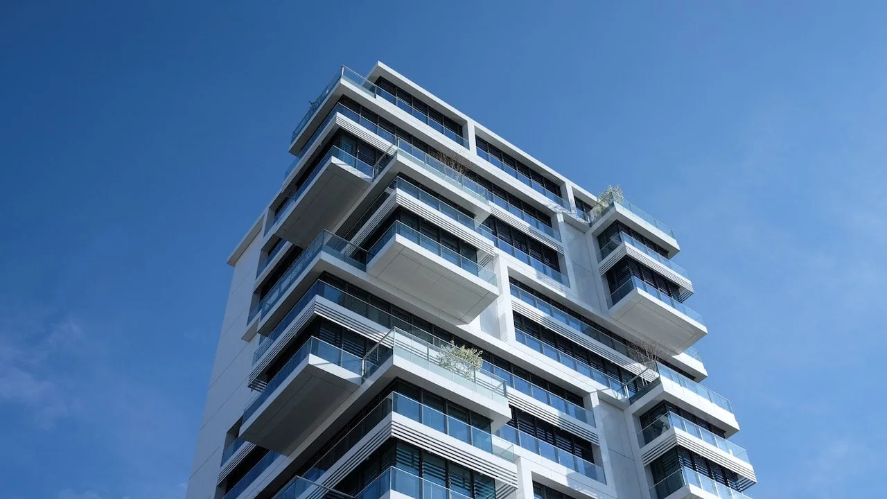 Edificio moderno de múltiples pisos con fachada blanca y balcones de cristal bajo un cielo azul claro. Representa inversión inmobiliaria y desarrollo urbano.