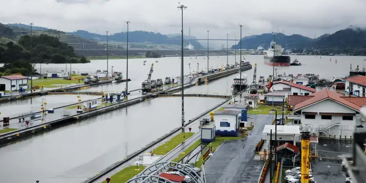 Vista de un sistema de esclusas con buques de carga, edificios operativos y grúas. Infraestructura clave para el comercio marítimo global, con montañas y cielo nublado de fondo.