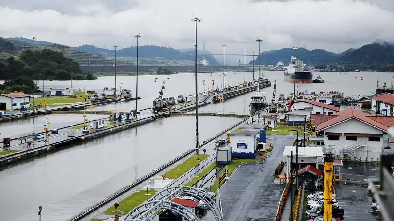 Vista de un sistema de esclusas con buques de carga, edificios operativos y grúas. Infraestructura clave para el comercio marítimo global, con montañas y cielo nublado de fondo.