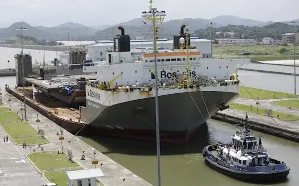 Gran buque de carga gris y blanco, con el nombre 'Boskalis', navegando por una esclusa de canal, asistido por un remolcador azul. Transporta una plataforma grande en su cubierta, con colinas verdes al fondo.