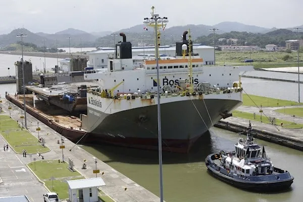 Gran buque de carga gris y blanco, con el nombre 'Boskalis', navegando por una esclusa de canal, asistido por un remolcador azul. Transporta una plataforma grande en su cubierta, con colinas verdes al fondo.