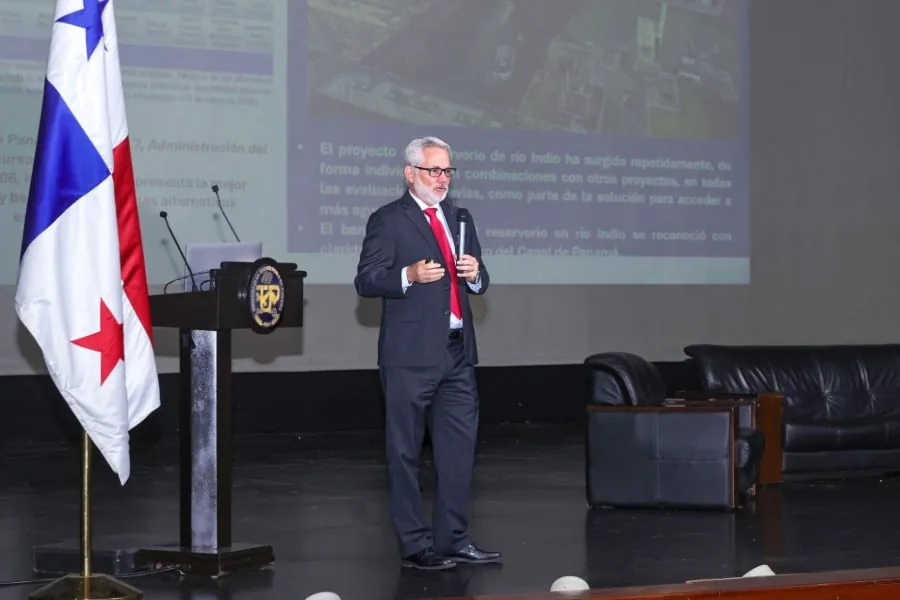Ponente en conferencia de negocios, con bandera panameña y pantalla mostrando datos de un proyecto, en un auditorio.