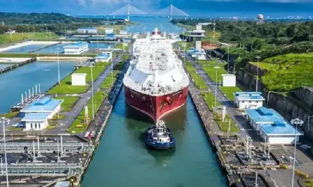 Vista aérea de un gran buque de carga, asistido por un remolcador, transitando por un sistema de esclusas de un canal. Se aprecian edificios de control, vegetación y un puente atirantado al fondo.