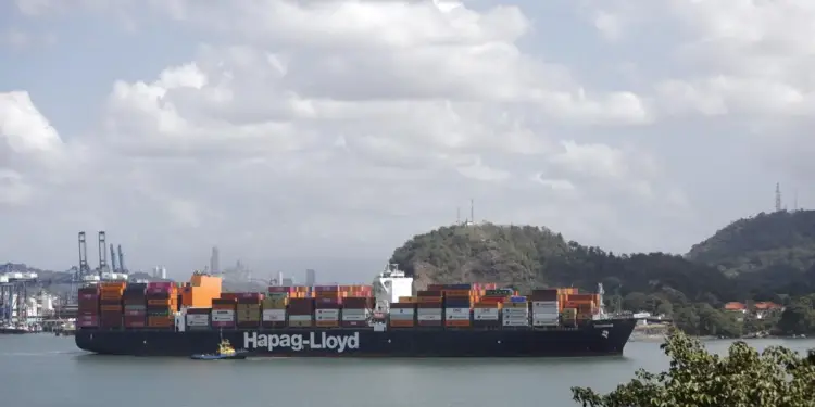 Un gran buque portacontenedores de Hapag-Lloyd navega por aguas tranquilas, asistido por un remolcador, con grúas portuarias y un horizonte urbano al fondo.