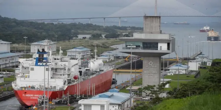Gran barco mercante navegando cerca de una torre de control y un puente colgante en un canal artificial.