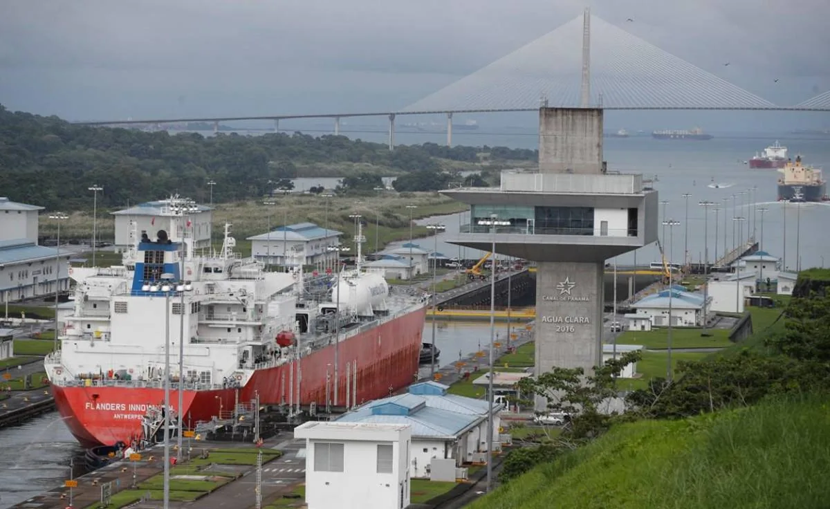 Gran barco mercante navegando cerca de una torre de control y un puente colgante en un canal artificial.
