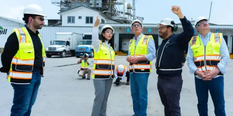 Grupo de profesionales con cascos y chalecos de seguridad inspeccionando una planta industrial con camiones de carga.