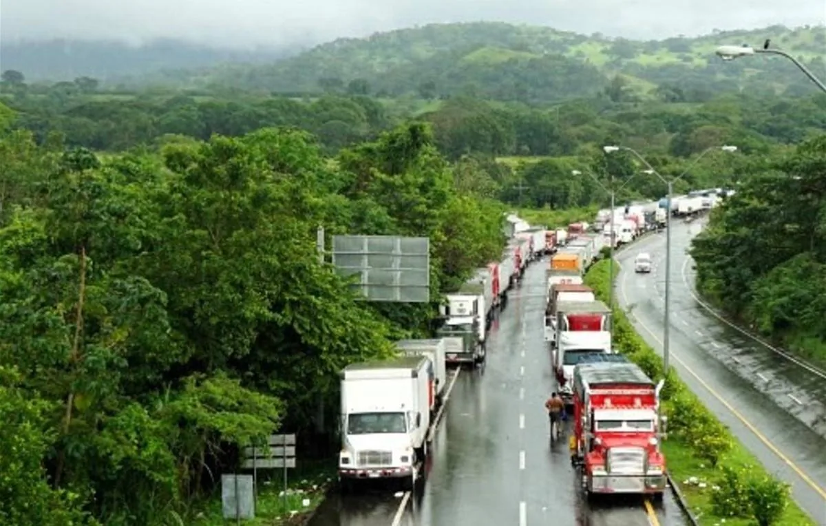 Vista aérea de una larga fila de camiones de carga detenidos en una carretera mojada, rodeada de densa vegetación y colinas verdes bajo un cielo nublado. Un hombre camina junto a un camión rojo.