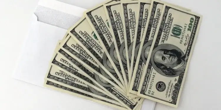 Abanico de billetes de cien dólares saliendo de un sobre blanco sobre fondo claro