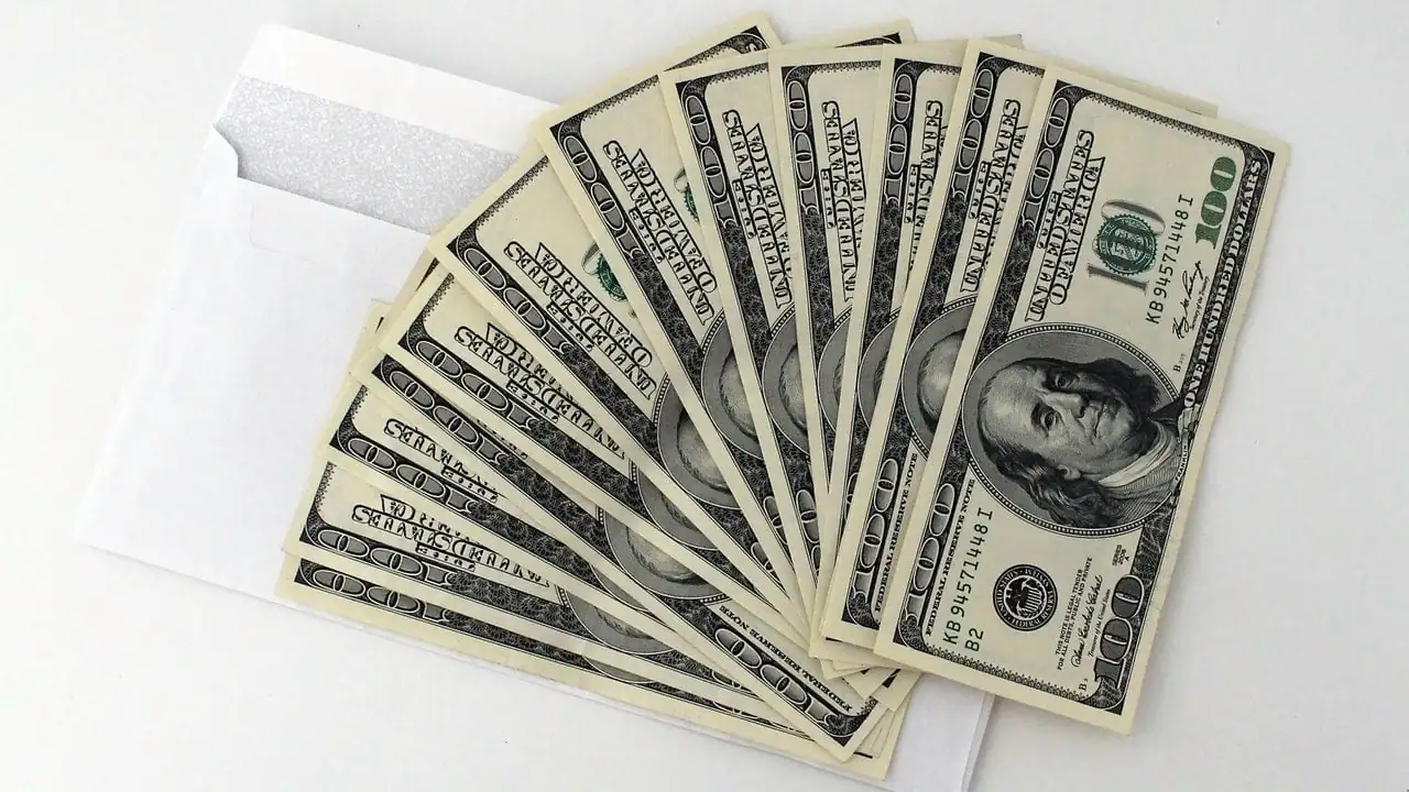 Abanico de billetes de cien dólares saliendo de un sobre blanco sobre fondo claro