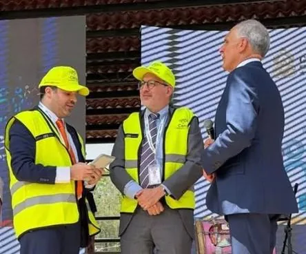 Tres profesionales en un escenario. Dos con chalecos y gorras amarillas; uno recibe un objeto. El tercero, de traje, sostiene un micrófono.