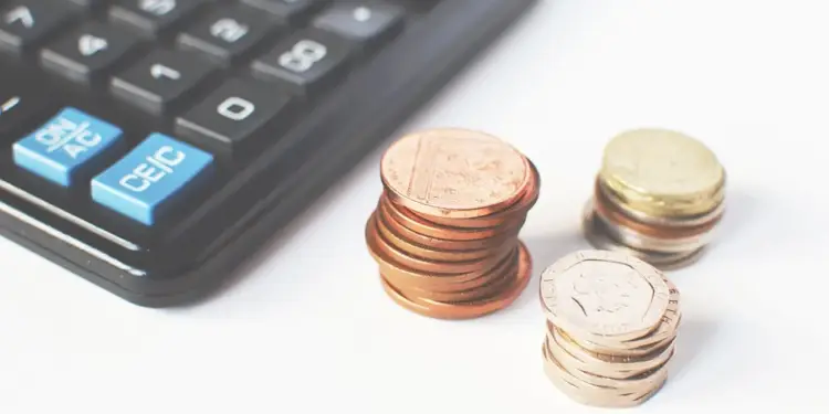 Una calculadora negra junto a tres pequeñas pilas de monedas sobre un fondo blanco.