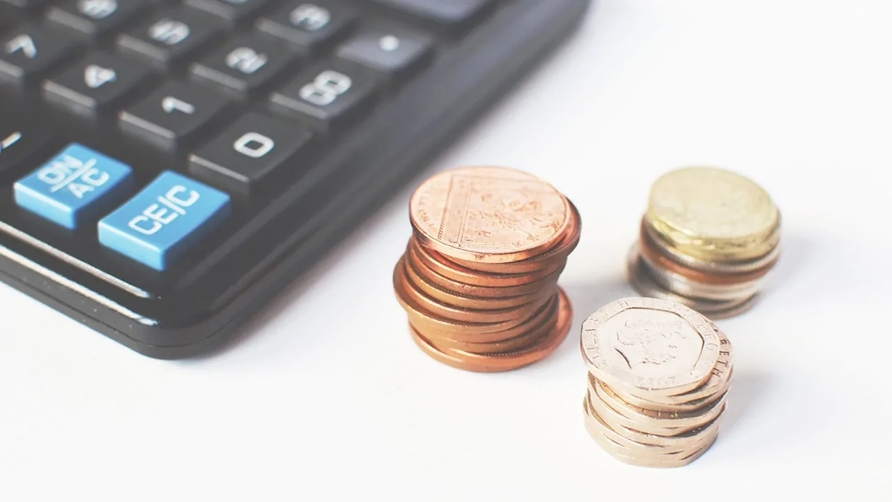 Una calculadora negra junto a tres pequeñas pilas de monedas sobre un fondo blanco.