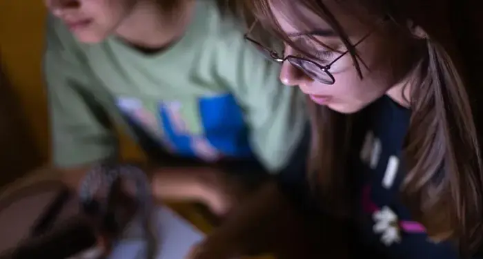 Dos niñas jóvenes iluminadas por la pantalla frente a un teclado blanco y una laptop en una habitación oscura