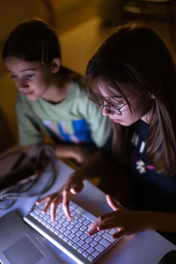 Dos niñas jóvenes iluminadas por la pantalla frente a un teclado blanco y una laptop en una habitación oscura