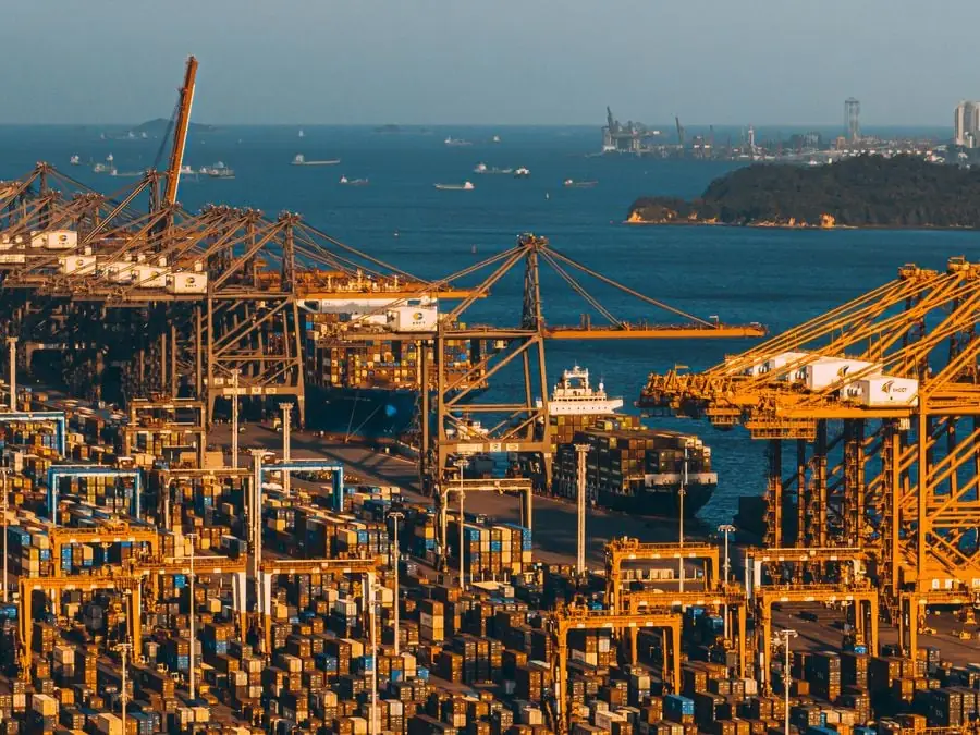 Vista aérea de un concurrido puerto con grúas gigantes y numerosos contenedores apilados junto al mar.