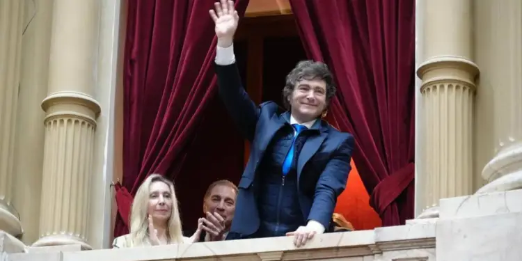 Hombre de traje azul saludando con la mano desde un balcón con cortinas rojas y columnas de piedra, acompañado por otras personas.