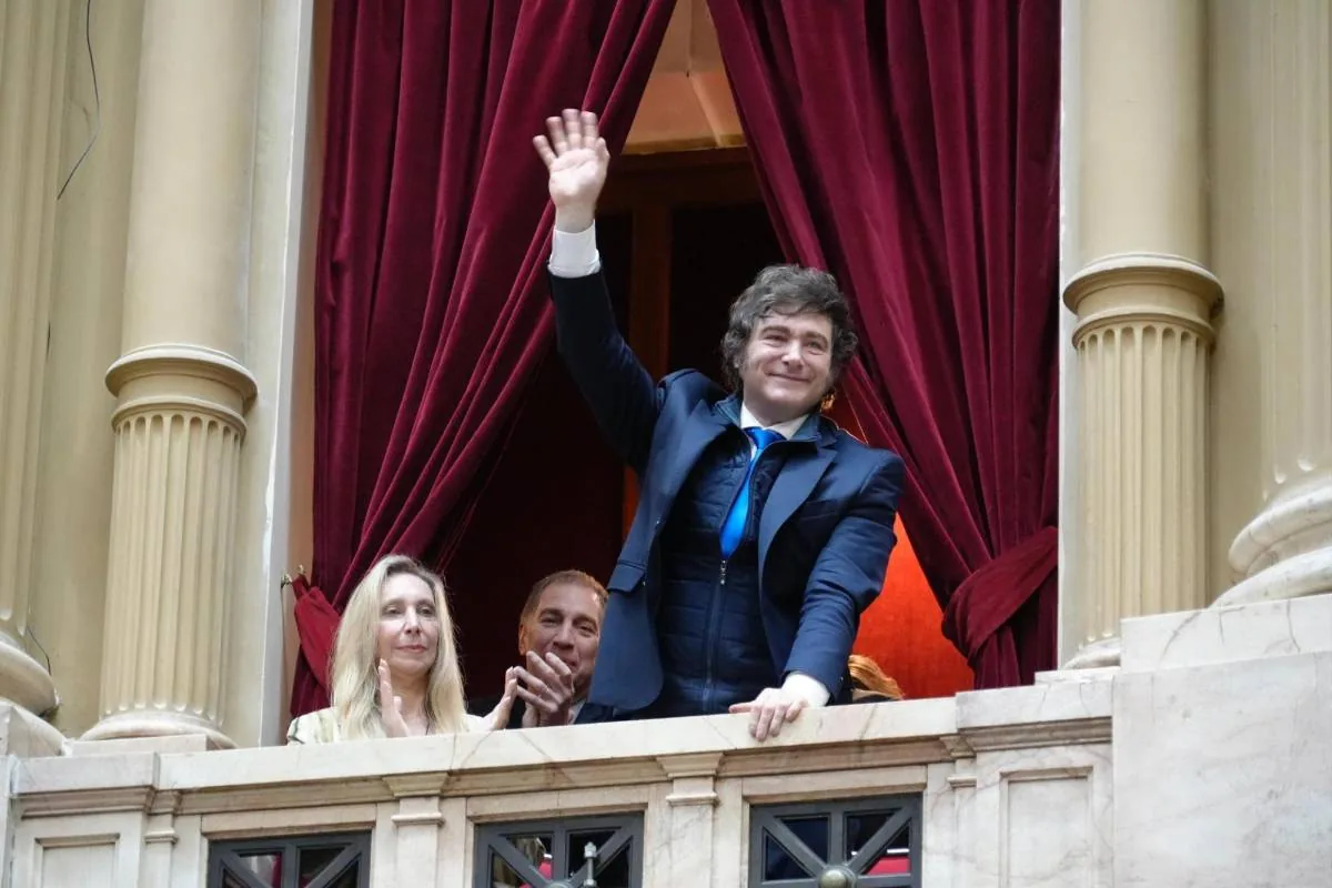 Hombre de traje azul saludando con la mano desde un balcón con cortinas rojas y columnas de piedra, acompañado por otras personas.
