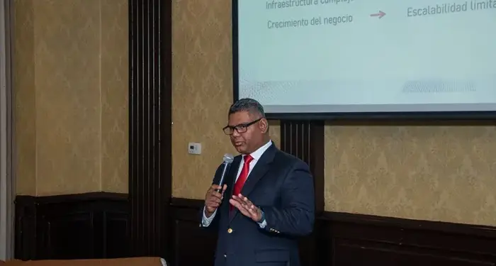 Un ponente con traje oscuro y corbata roja hablando ante una pantalla de proyección con texto en una sala de conferencias.