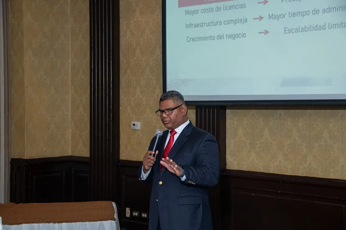 Un ponente con traje oscuro y corbata roja hablando ante una pantalla de proyección con texto en una sala de conferencias.