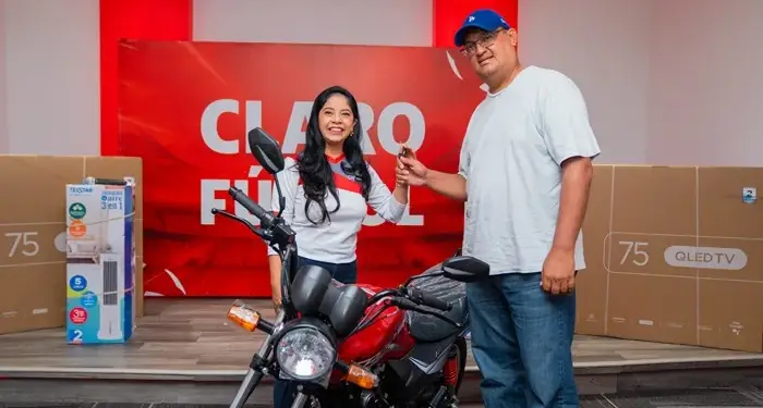 Una mujer entrega las llaves de una motocicleta roja a un hombre junto a cajas de electrodomésticos en un evento