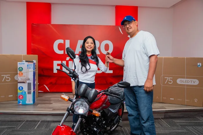 Una mujer entrega las llaves de una motocicleta roja a un hombre junto a cajas de electrodomésticos en un evento