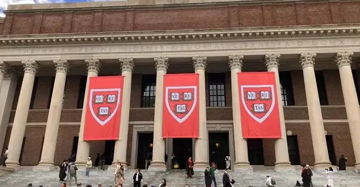 Fachada de la Biblioteca Widener de Harvard con columnas clásicas y tres estandartes rojos con el escudo 'VE RI TAS'. Numerosas personas suben y bajan las escaleras.