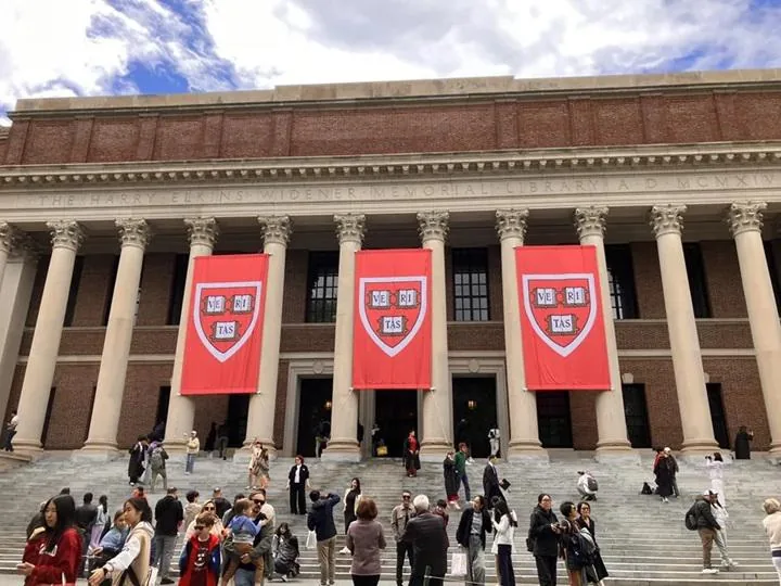 Fachada de la Biblioteca Widener de Harvard con columnas clásicas y tres estandartes rojos con el escudo 'VE RI TAS'. Numerosas personas suben y bajan las escaleras.