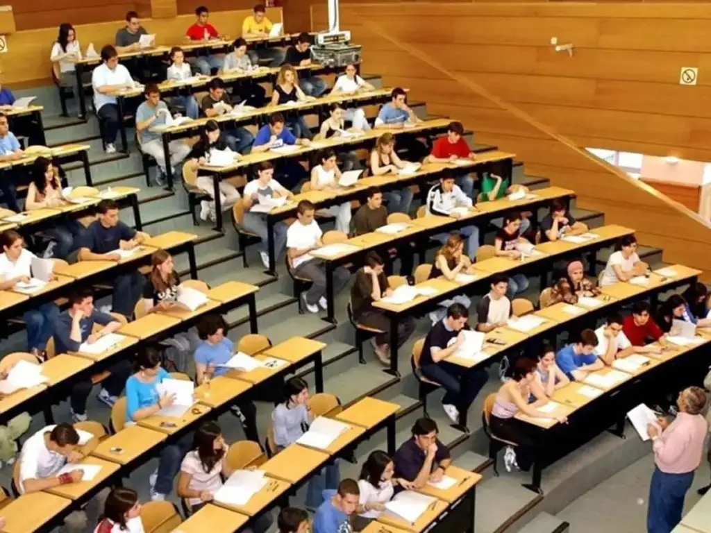 Vista aérea de un aula magna llena de estudiantes sentados en pupitres, prestando atención a un instructor al frente.