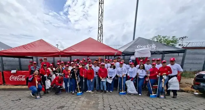 Grupo de voluntarios con gorras rojas, escobas y palas frente a carpas de Coca-Cola en jornada ambiental.