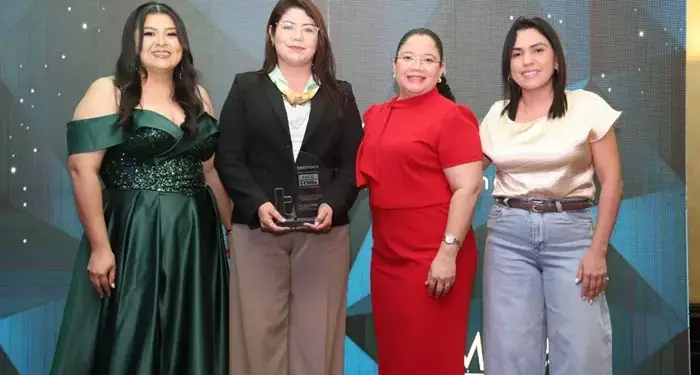 Cuatro mujeres profesionales sonriendo y posando de pie frente a un fondo decorado mientras una de ellas sostiene un reconocimiento.