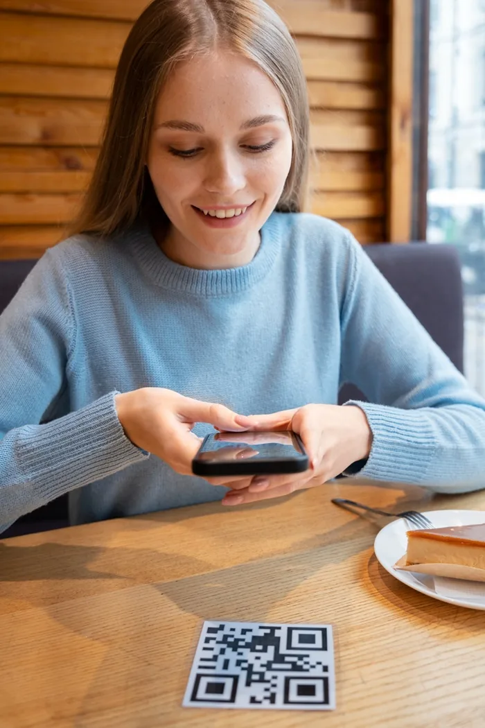 Mujer joven sonriente escaneando un código QR en la mesa con su teléfono móvil en un establecimiento