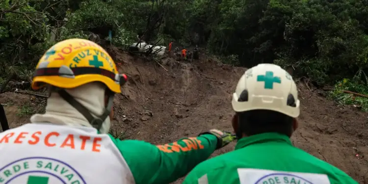 Dos rescatistas de espaldas con cascos y uniformes observan una zona afectada por un deslizamiento de tierra en la montaña.