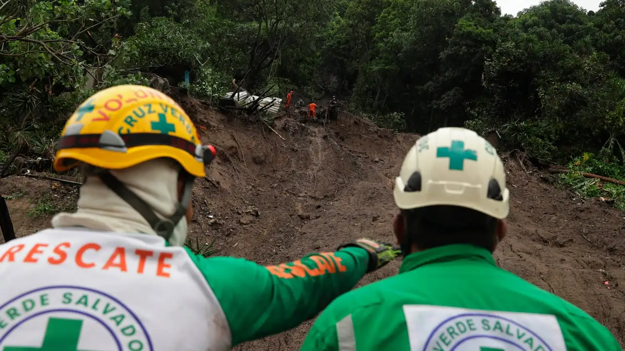 Dos rescatistas de espaldas con cascos y uniformes observan una zona afectada por un deslizamiento de tierra en la montaña.