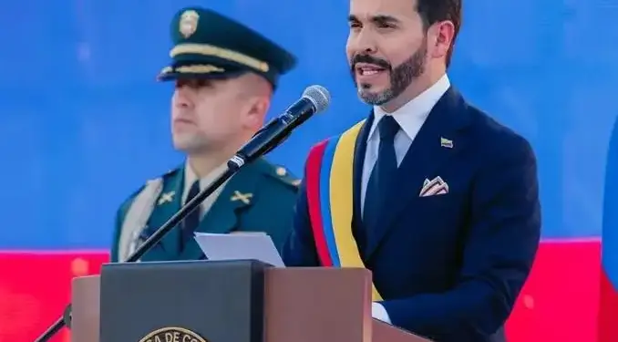 Hombre con traje y banda presidencial hablando en un atril, con un oficial de policía al fondo.