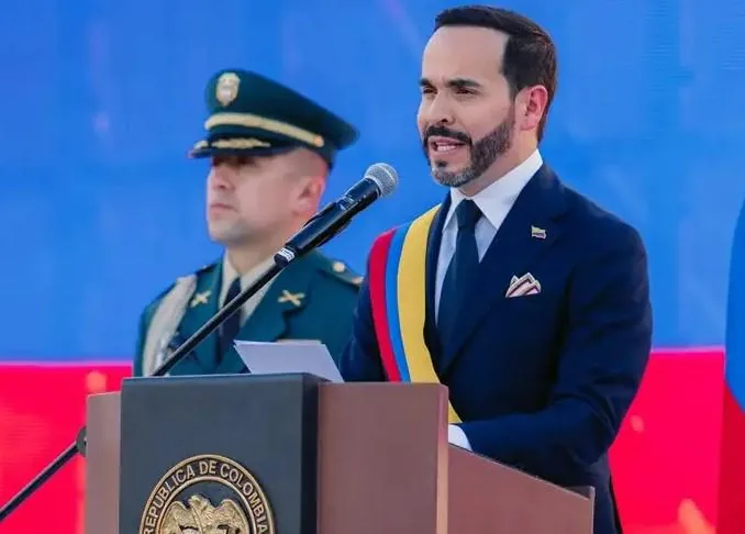 Hombre con traje y banda presidencial hablando en un atril, con un oficial de policía al fondo.