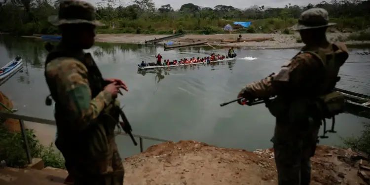 Dos soldados armados vigilan un bote con numerosas personas navegando por un río caudaloso.