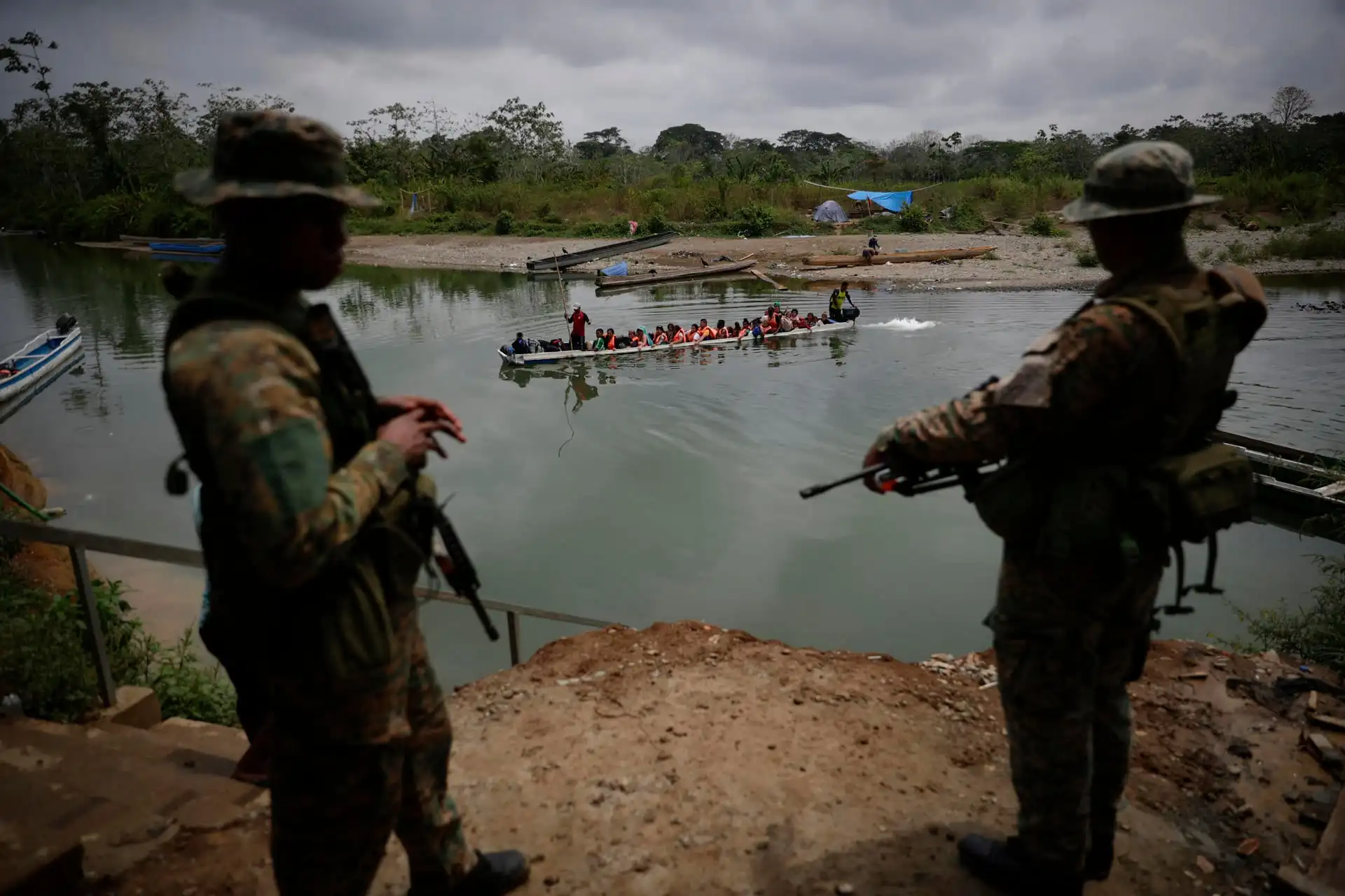 Dos soldados armados vigilan un bote con numerosas personas navegando por un río caudaloso.