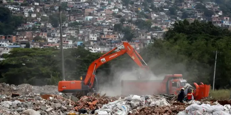 Excavadora naranja cargando escombros en un camión con una zona urbana densamente poblada al fondo