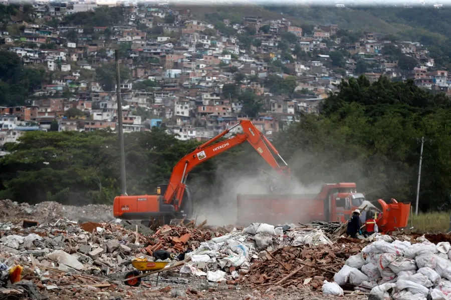 Excavadora naranja cargando escombros en un camión con una zona urbana densamente poblada al fondo
