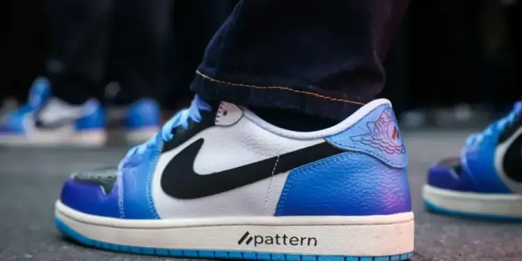 Primer plano de una zapatilla deportiva blanca y azul con detalles negros, suela blanca y el texto 'pattern'. Se aprecian jeans oscuros y un logo alado en el talón, con otras zapatillas similares al fondo.