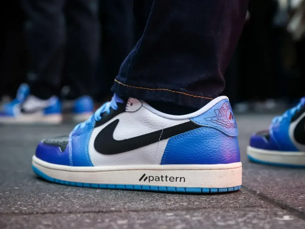 Primer plano de una zapatilla deportiva blanca y azul con detalles negros, suela blanca y el texto 'pattern'. Se aprecian jeans oscuros y un logo alado en el talón, con otras zapatillas similares al fondo.