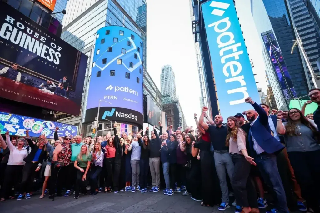 Gran grupo de personas celebrando con puños en alto frente a pantallas gigantes de Nasdaq y Pattern, simbolizando un éxito empresarial.