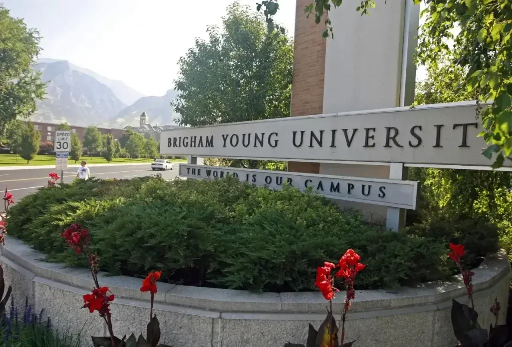 Entrada a Brigham Young University con su letrero principal, arbustos verdes, flores rojas y montañas al fondo. Un coche y una persona en la carretera.