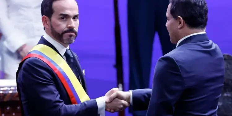 Dos hombres en traje formal se dan la mano. El de la izquierda lleva una banda tricolor (amarillo, azul, rojo). Simboliza un acuerdo o colaboración profesional en un evento.