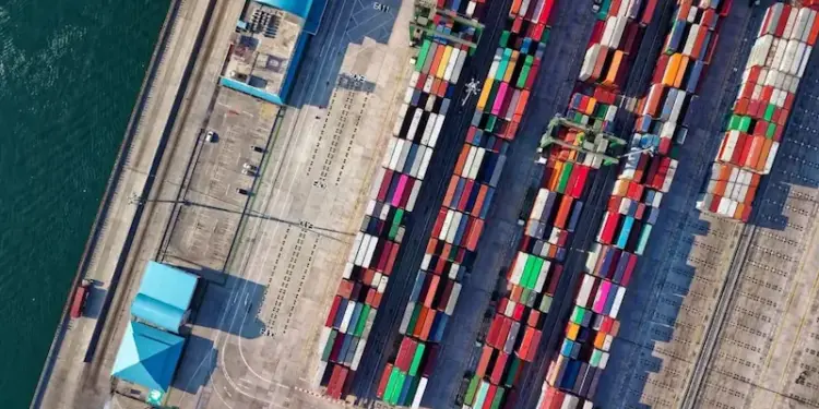 Vista aérea de un puerto comercial con múltiples contenedores de carga de colores apilados. Se observan grúas, muelles y agua.