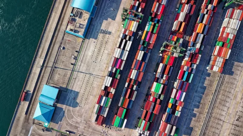 Vista aérea de un puerto comercial con múltiples contenedores de carga de colores apilados. Se observan grúas, muelles y agua.