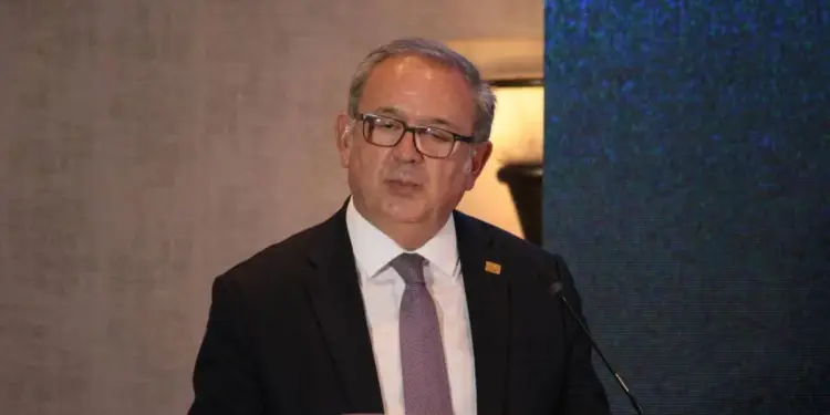 Hombre de mediana edad con traje oscuro, corbata y gafas, hablando en un podio blanco con micrófono. Fondo con pared texturizada y pantalla digital azul.