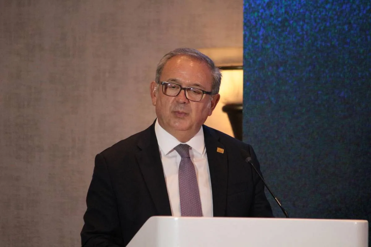 Hombre de mediana edad con traje oscuro, corbata y gafas, hablando en un podio blanco con micrófono. Fondo con pared texturizada y pantalla digital azul.